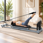 Woman lying on a grey foldable carbon-steel Pilates reformer with her feet in the pulley straps in a home studio